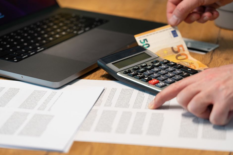 Mains utilisant une calculatrice à côté d’un billet de cinquante euros, d’un ordinateur portable et de documents